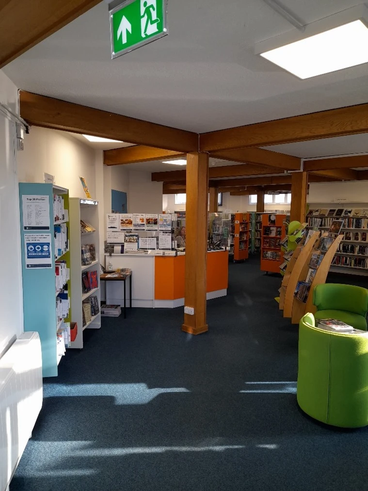 This picture has a white and orange front desk with a library assistant sitting behind it.  There are shelves of books to the left, right and back of the picture.