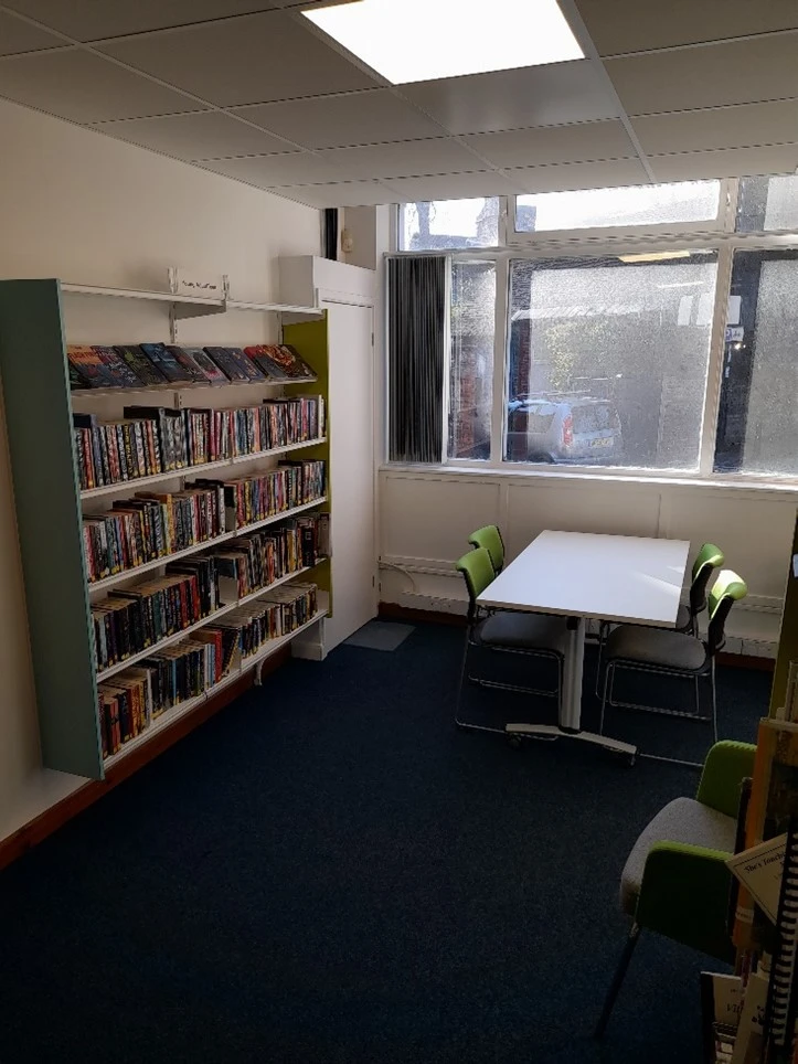 This picture has a book shelf to the left filled with books and a white table with four green chairs to the right.