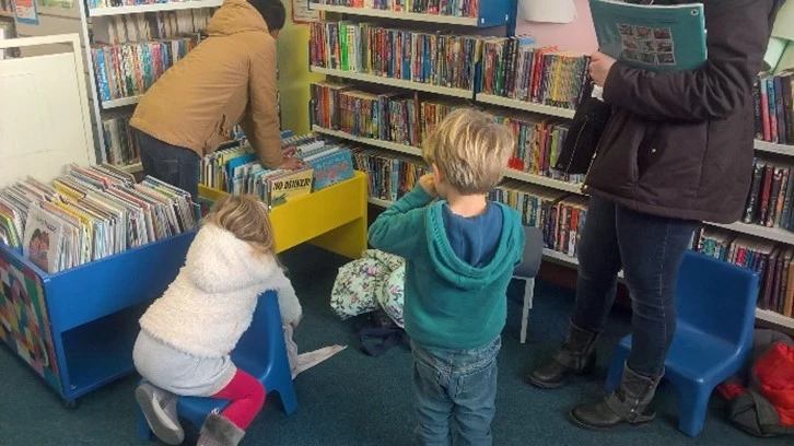This picture shows three children looking at the books in the children's section and an adult standing to the right of them.