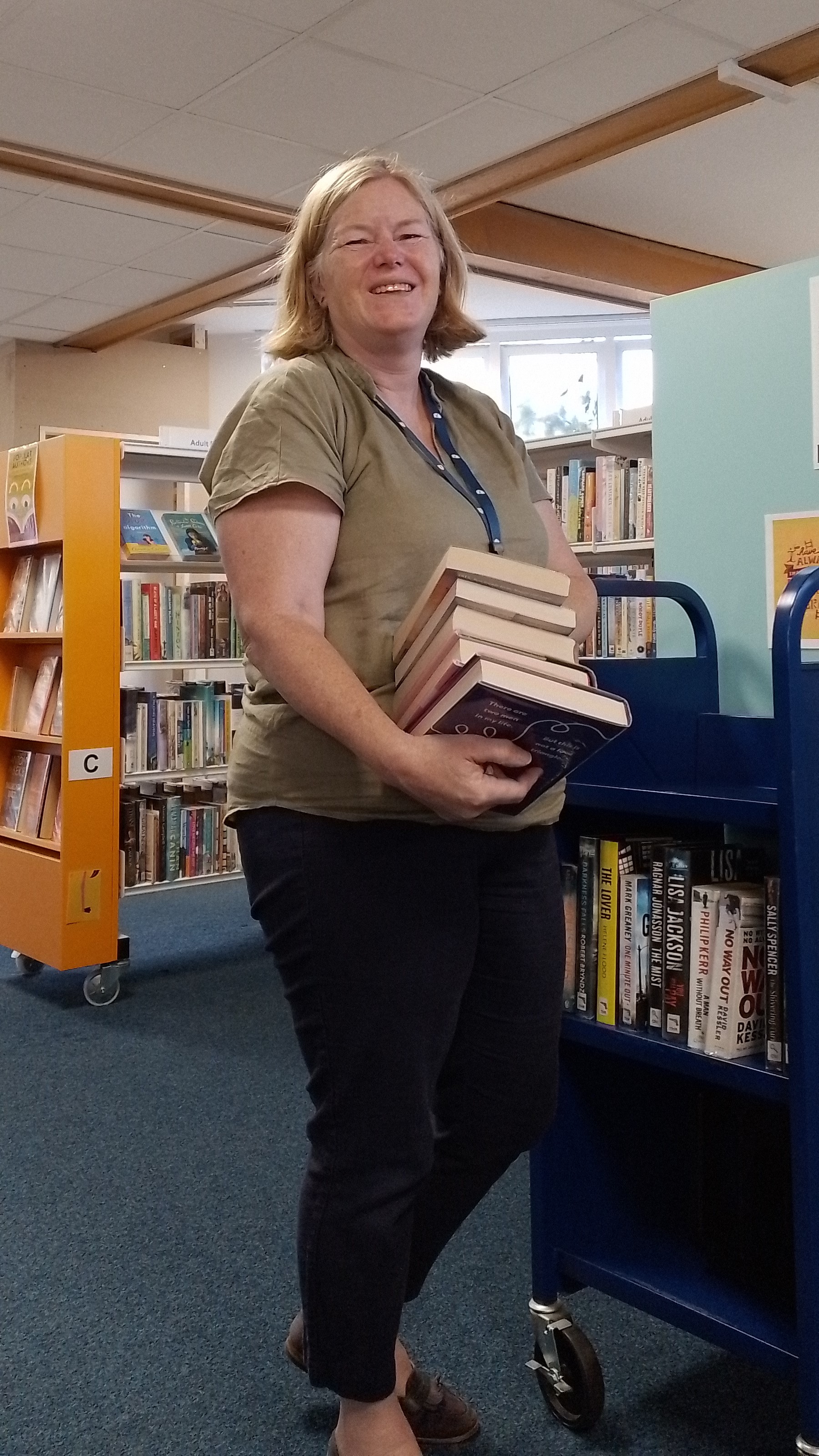 This is a picture of a library assistant holding a stack of books, standing and smiling at the camera.  There are shelves of books behind them.