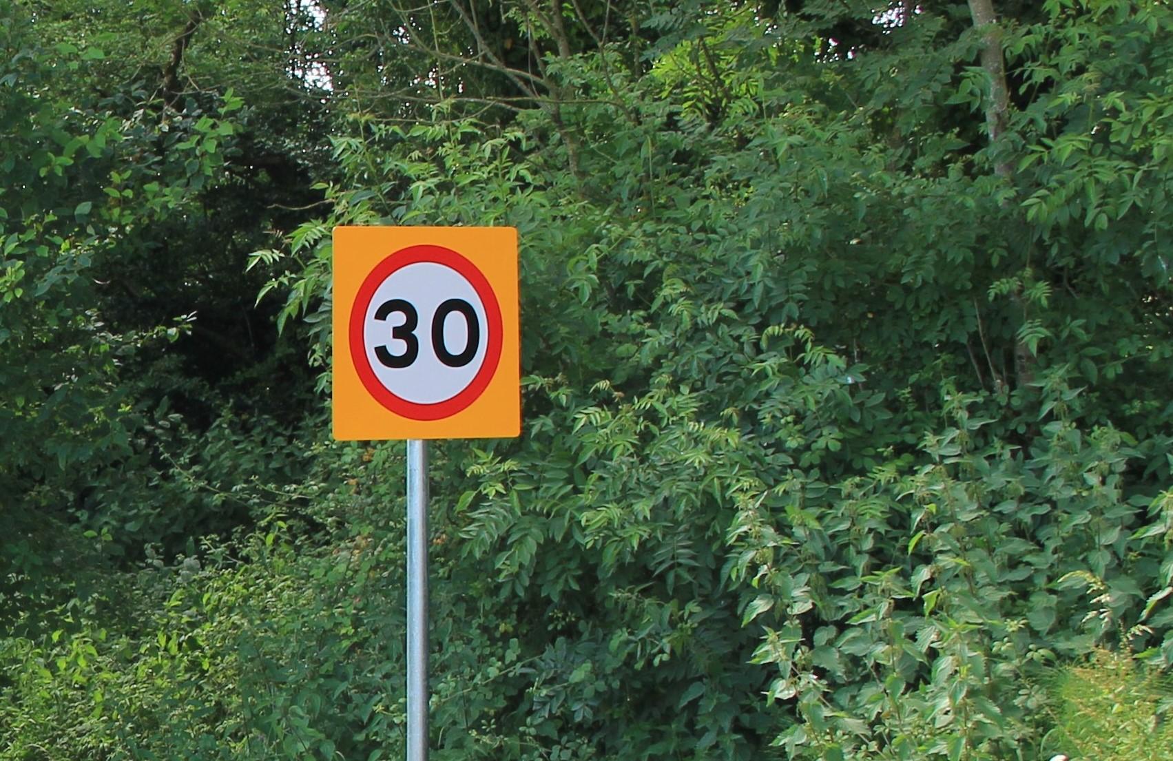 A 30mph sign on a Dorset road on a yellow background with hedgerow behind