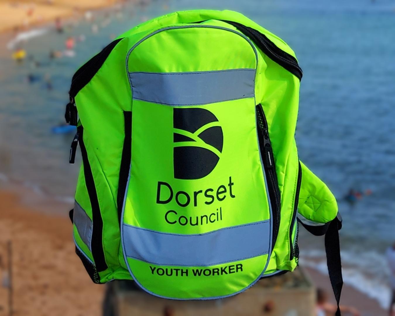 A yellow backpack sits on a fence at West Bay in Dorset. The text on the backpack read Dorset Council Youth Worker and has the Dorset Council logo on it.