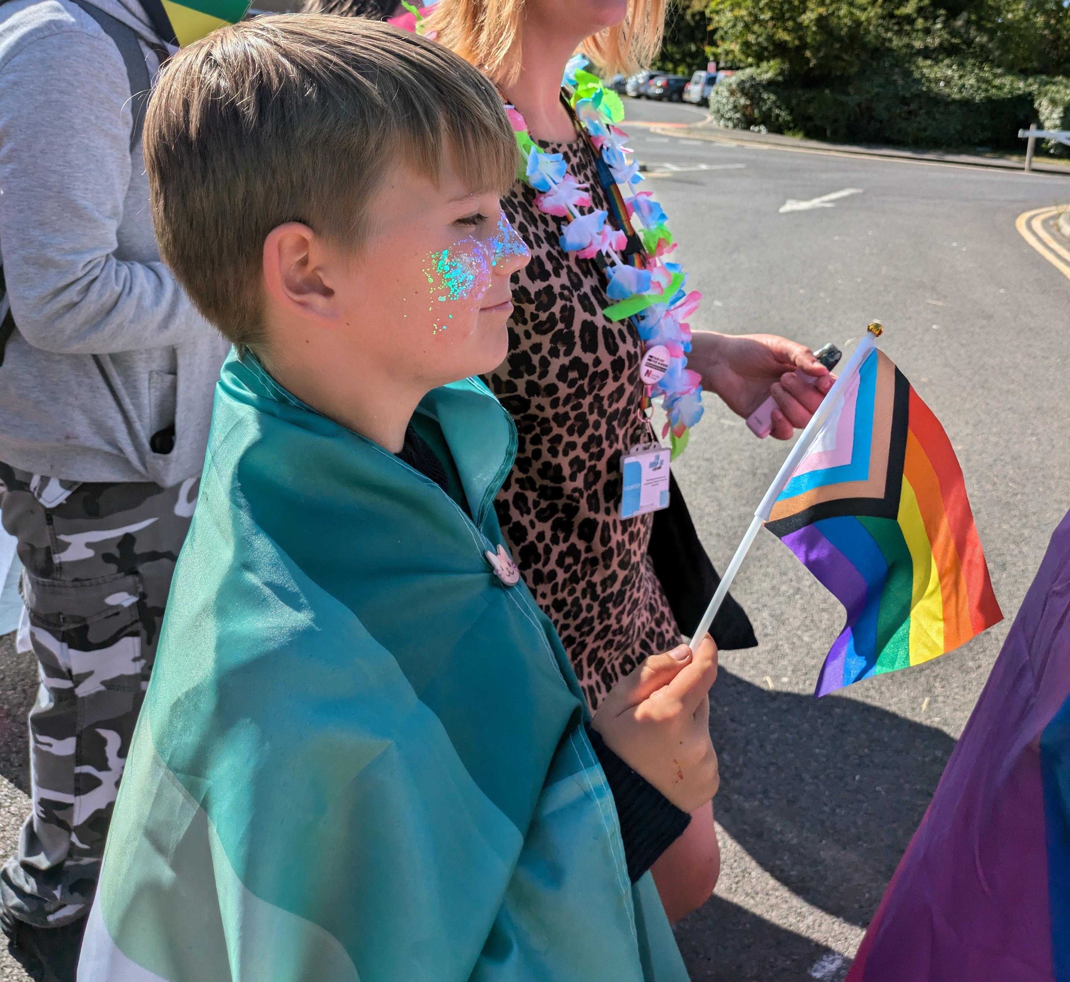 A boy is smiling and holding an LGBTQ+ flag. He also has glitter on his face and a flag draped around his shoulders.