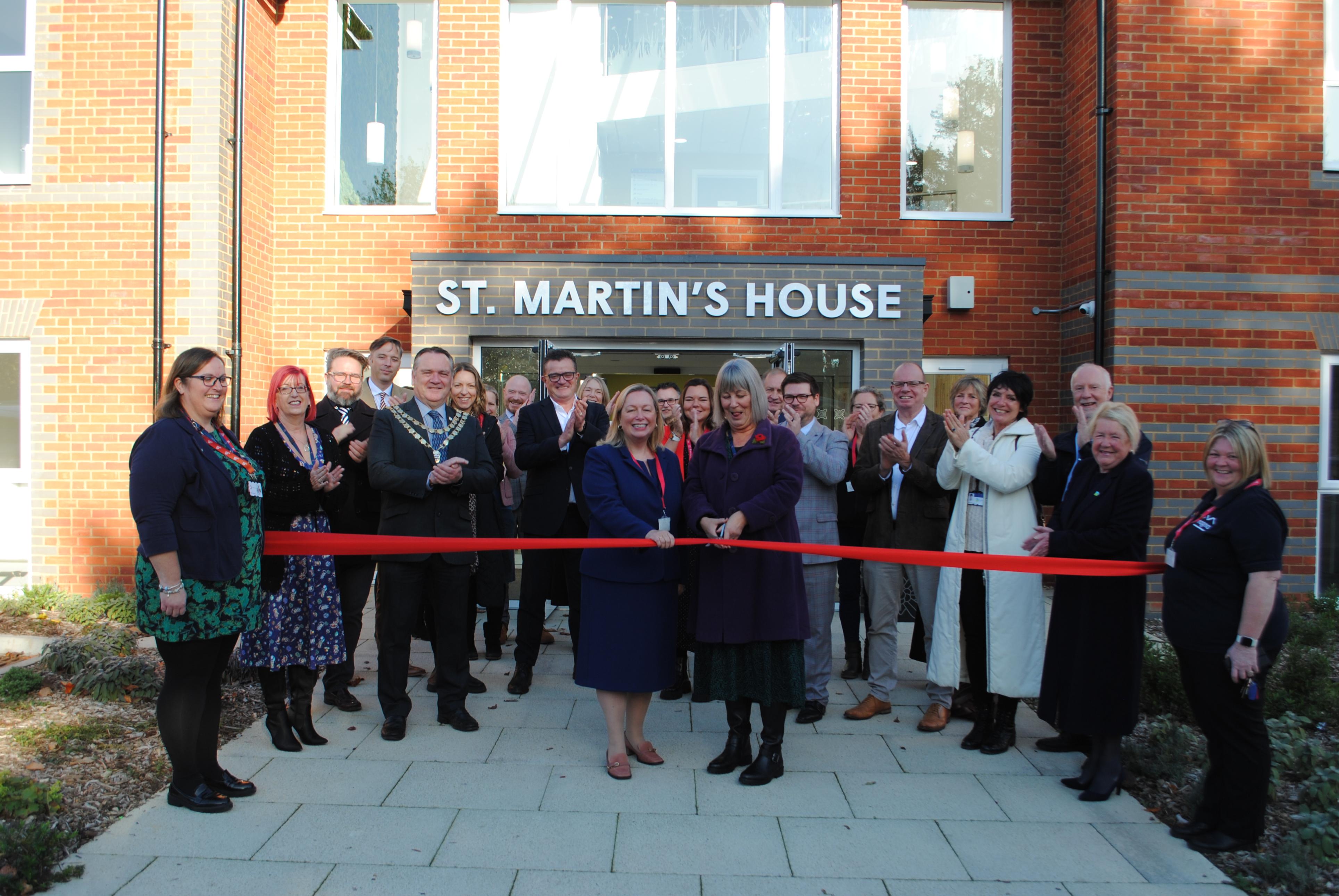 Over 20 people are stood together and cheering outside the entrance of a red brick large building with the name of the building - St Martin&#39;s House - above the large glass doors. Two women are stood in the middle and are about to cut a red ribbon.