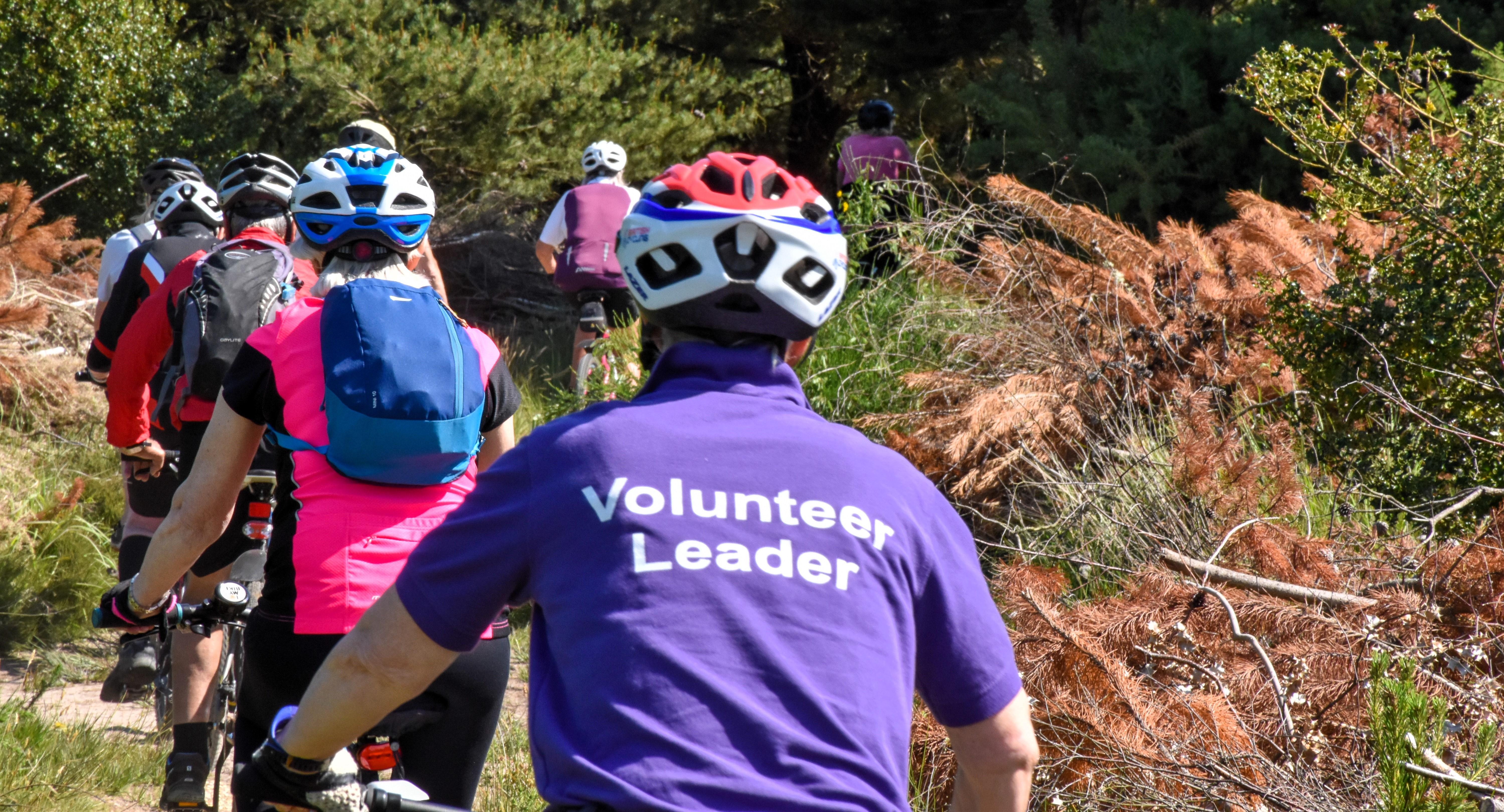 group of individuals ride bikes on trail through woods, the person at the back has a t-shirt on that reads &#39;volunteer leader&#39;