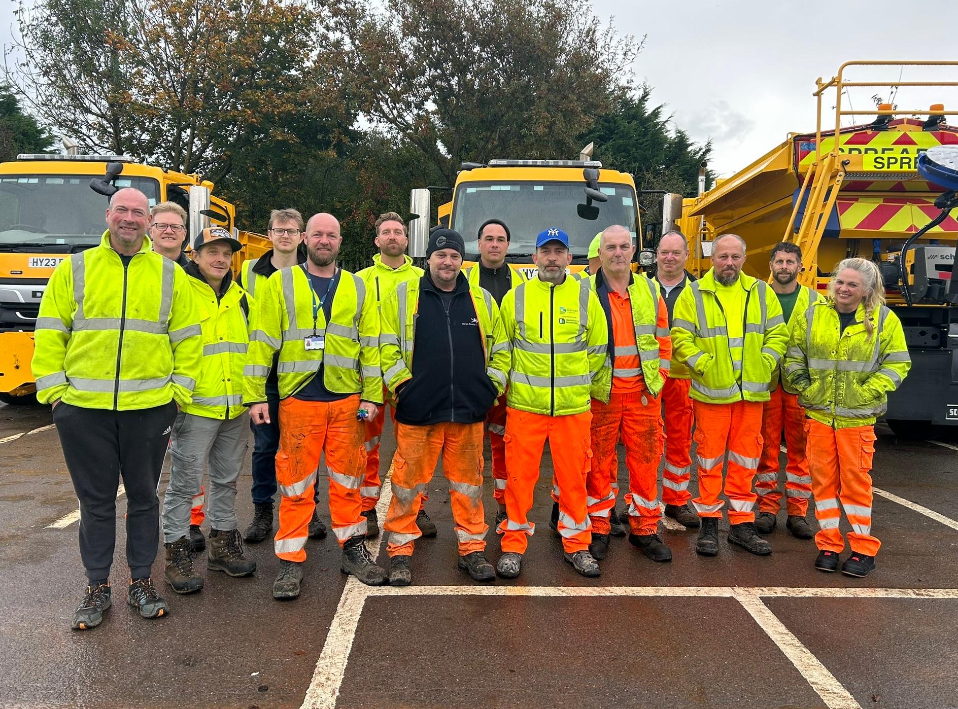 People standing in front of a gritter lorry. They are wearing yellow jackets and orange trousers.