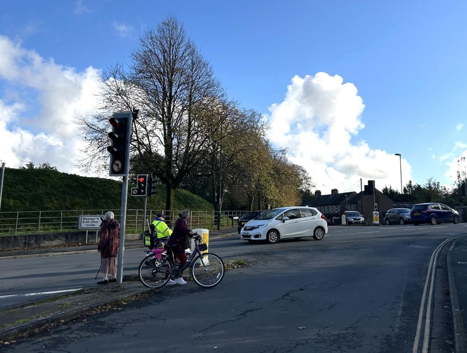 Three people waiting to cross a busy junction with vehicles passing by