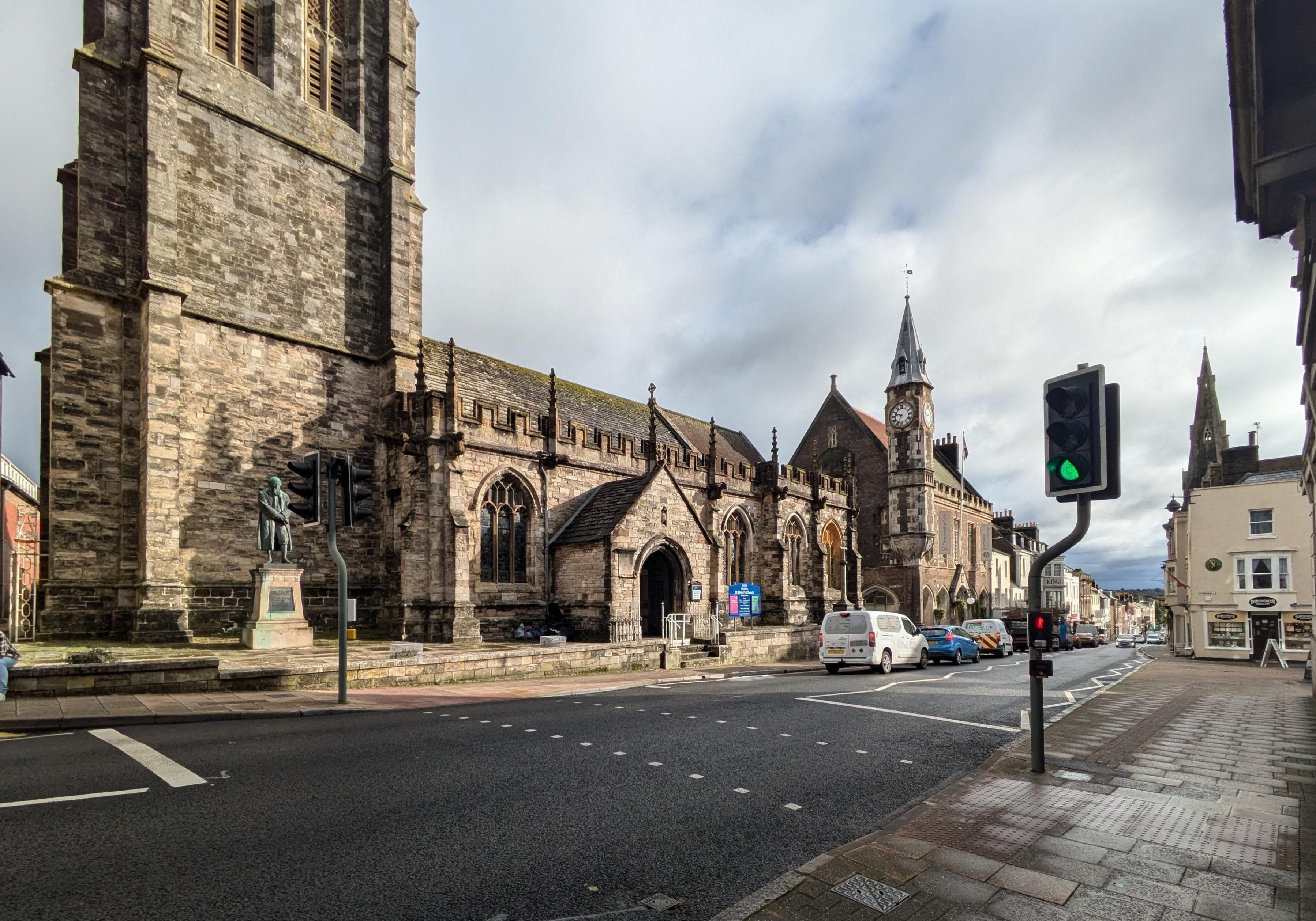 A pedestrian crossing with a church and clock tower in the background and a line of vans and cars driving away from the crossing