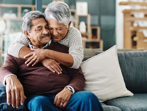 Two people, one sat on a sofa and the other leaning over the back cuddling them from behind