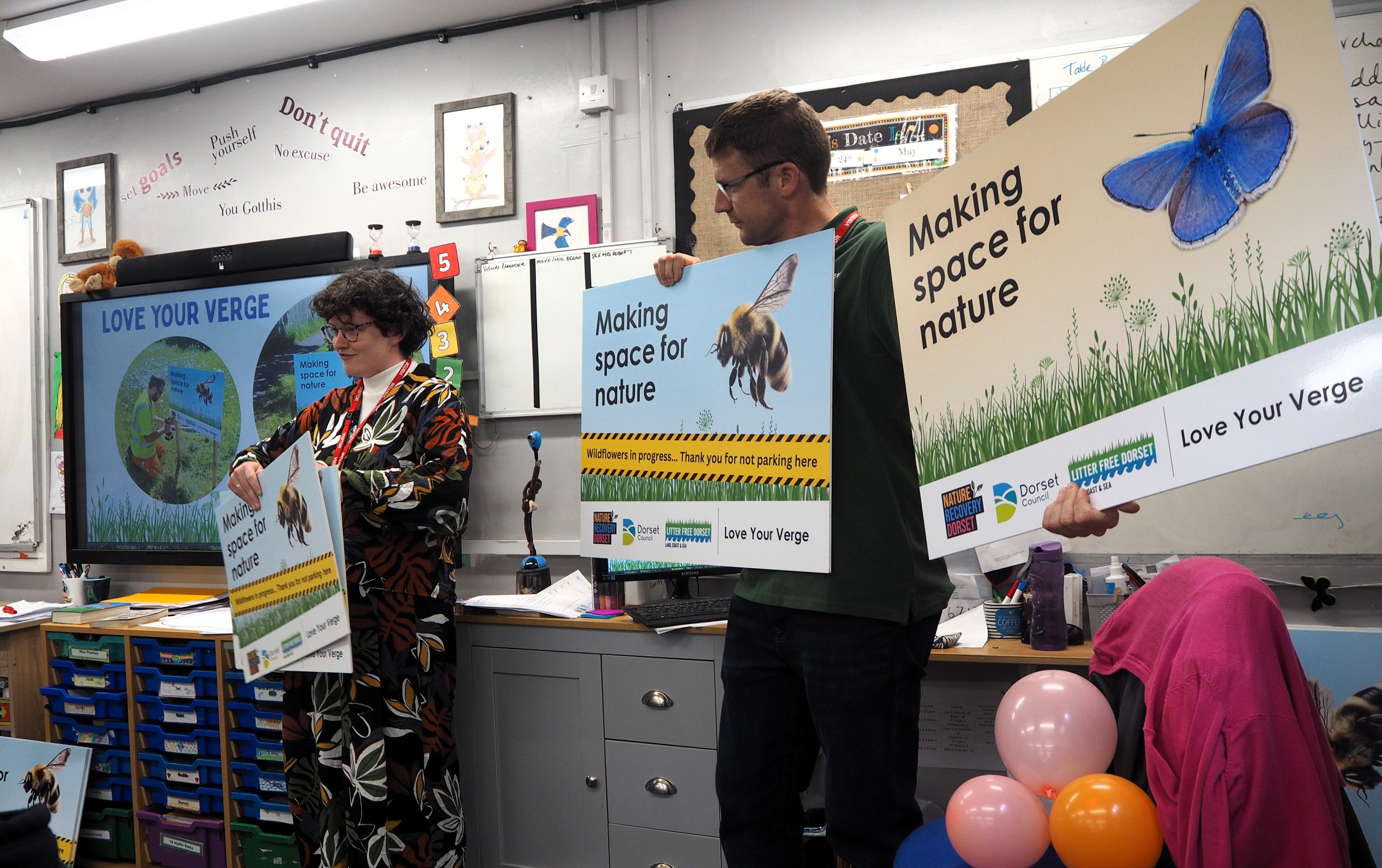 two council employees hold up 'making space for nature' signs at the front of a classroom