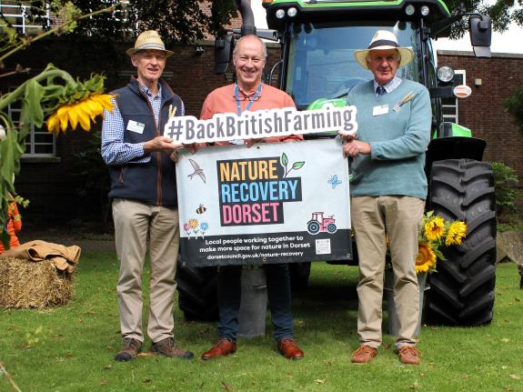 three individuals pose for picture in front of tractor holding 'Nature Recovery Dorset' and 'Back British Farming' signs