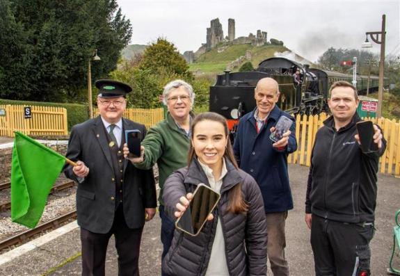 Five people picture on a railway platform with a steam train and Corfe Castle in the background. There are four men, one of whom is dressed in railway uniform and holding a green flag, plus a woman with long brown hair.