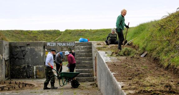 People help clear vegetation using wheelbarrows and rakes at historic site, graffiti is visible on the walls of the ruins