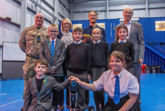 6 children in uniform pose for a photograph with a small segway, 4 adults stand in the background of the photo, one blonde woman in glasses, one man uniform and two smartly dresses men, one of which holds a plasticine boat.