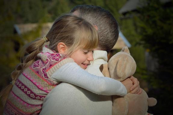 a little girl is hugging her foster parent tightly. She is also holding a soft toy dog.