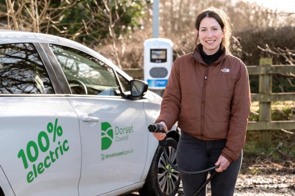 individual poses holding an electric vehicle charging lead in front of a white electric vehicle. An electric vehicle charger can be seen in the background. And the car has 100% electric and the Dorset Council logo on its side.