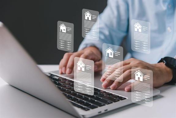 A white man is using a silver laptop. You can see his hands resting on the base of the open laptop. Graphics of houses appear to popping up from the keyboard.