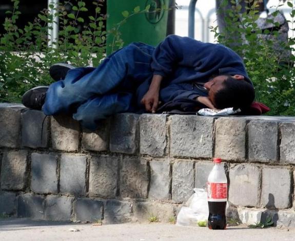 A homeless man who is wearing blue jeans and a blue jumper is laid on his left side on a grey stone brick wall